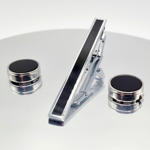 Puede incluir: Un conjunto de accesorios formales, que incluye un clip de corbata plateado con una franja central negra y dos gemelos redondos a juego con centros negros. Los artículos se muestran sobre una superficie reflectante.