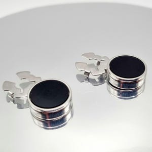 May include: A pair of silver cufflinks with a circular black centre. The cufflinks have a shiny, metallic finish and a unique, stylized design. The cufflinks are displayed on a reflective surface.