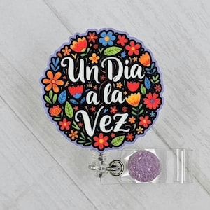 May include: A round badge reel with a floral design and the text "Un Dia a la Vez" in white. The flowers are in shades of red, orange, yellow, and blue. The badge reel has a clear plastic holder and a purple, glittery accent.