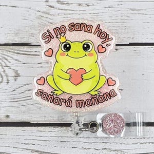 May include: A light green frog badge with a crown, holding a red heart, and the Spanish text "Si no sana hoy, sanará mañana" on a pink glitter background. Includes a retractable reel with a pink glitter accent.