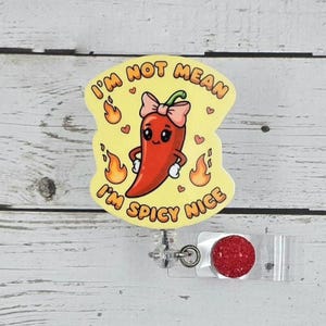 May include: A yellow badge reel with a cartoon chili pepper design. The red chili pepper has a pink bow and is surrounded by flames and hearts. Text on the badge reads "I'm not mean, I'm spicy nice."