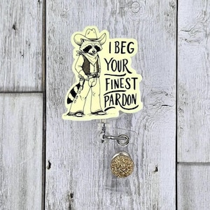 May include: A badge reel featuring a cartoon raccoon dressed as a cowboy, with the text "I BEG YOUR FINEST PARDON." The reel has a clear plastic body, a metal clip, and a gold glitter button.