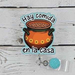 May include: A badge reel with a cartoon image of a pot of beans with the text "Hay comida en la casa". The reel has a turquoise glitter accent.