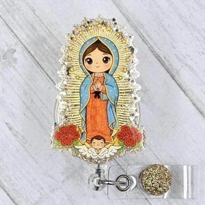 May include: A badge reel featuring a cartoon illustration of Our Lady of Guadalupe. The image shows a figure in a blue cloak and orange dress, surrounded by a gold halo with stars. Red roses and an angel are at the bottom. The reel has a glittery gold button.