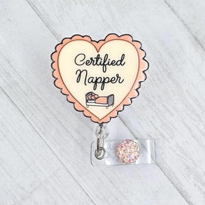 May include: A heart-shaped badge reel with a scalloped edge, featuring the words "Certified Napper" and an illustration of a bed. The badge reel has a clear plastic clip and a decorative, sparkly pink button.
