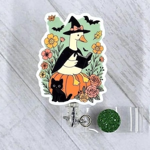 May include: A Halloween-themed badge reel featuring a cartoon goose dressed as a witch, reading a book while sitting on a pumpkin. A black cat sits nearby. The design includes flowers, bats, and leaves. The reel has a green glitter button.