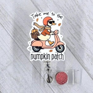 May include: A badge reel with a cartoon duck riding a pink scooter, wearing a helmet, sunglasses, and a scarf. The duck carries a pumpkin in a basket. Text reads "Take me to the pumpkin patch."