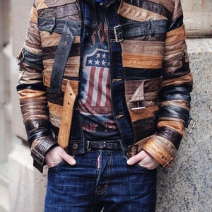 May include: A brown, tan, and black leather patchwork jacket with horizontal panels and metal buckles. The jacket is worn with a graphic tee and blue jeans.