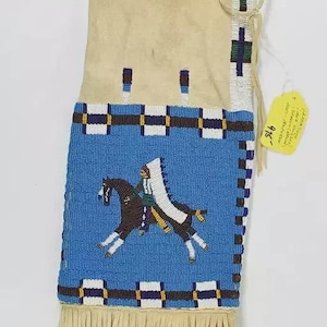 Puede incluir: Una bolsa de cuero color canela con un panel de cuentas azul que representa a un nativo americano a caballo. La bolsa tiene un fondo de flecos y detalles de cuentas en azul, blanco, verde y amarillo. Una etiqueta está adjunta.
