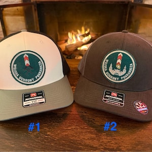 May include: Two baseball caps with a mesh back and a curved brim. One cap is white and gray, the other is dark gray. Both caps feature a circular patch with Polish text and a border post design.