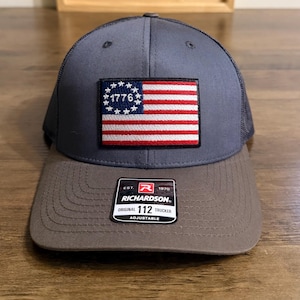 May include: A charcoal grey trucker hat featuring a rectangular patch of the Betsy Ross American flag with the year 1776 in the blue field. The hat has a solid front panel, a mesh back, and a sticker on the brim reading Richardson 112 Trucker.