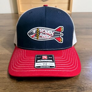 May include: A navy blue and white trucker hat with a red brim and white stitching. The front features an embroidered patch of a grey bomb with a shark face and the text BOMBS AWAY. A black and white sticker on the brim reads RICHARDSON 112.