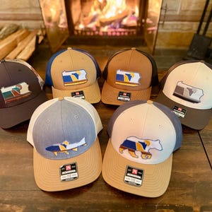 May include: A collection of trucker hats with various designs. The hats feature a mesh back and a curved brim. Designs include a fish, a bear, and a state outline. The hats are in shades of brown, blue, and tan.