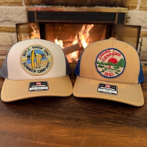 May include: Two trucker hats with embroidered patches. One hat has a patch with text "Rifle River DAC Summer Camp 1971". The other hat has a patch with text "Spring Camporee 1961". Both hats have a tan brim and a Richardson 112 tag.