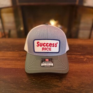 Peut inclure: Casquette de camionneur grise et blanche avec une visière verte. Le chapeau présente un écusson rectangulaire avec les mots "Success Rice" en lettres rouges. C'est une Richardson 112 Trucker.