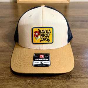 May include: A trucker hat featuring a cream-colored front panel, a tan brim, and dark navy mesh back panels. A rectangular yellow patch on the front displays a red flower and the text HAVE A NICE DAY. A black Richardson 112 sticker is on the brim.