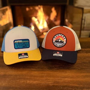 May include: Two trucker hats with mesh backs sit on a wooden surface. One hat is white, blue, and yellow with a patch that reads "Montana Big Sky Country". The other is red, white, and navy blue with a mountain patch.