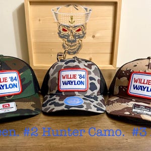 May include: Three baseball caps featuring a rectangular patch with the text WILLIE '84 WAYLON in red and blue. The left cap is green camouflage, the center is grey camouflage, and the right is brown camouflage. Each cap has a black mesh back.