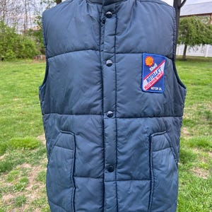 May include: A navy blue quilted vest with a high collar and button closure. The vest features two front pockets and a Shell Rotella Motor Oil patch. The vest is displayed on a mannequin against a backdrop of green grass and trees.