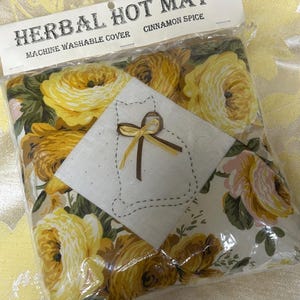 Pode incluir: Um tapete quente de ervas com padrão floral e capa lavável na máquina. O tapete apresenta um desenho de rosas amarelas e douradas. Um quadrado branco com um contorno de gato e um laço é costurado na frente. A embalagem diz "Herbal Hot Mat" e "Cinnamon Spice."