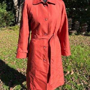 May include: A solid burnt orange trench coat featuring a classic collar, long sleeves with adjustable cuff straps, and a matching tie belt at the waist. The coat is secured with a front button closure and reaches below the knee in length.