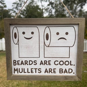 May include: A rectangular wooden sign with a light brown frame. The sign features two cartoon toilet paper rolls, one smiling and one frowning. Below the images, the text reads "BEARDS ARE COOL. MULLETS ARE BAD."