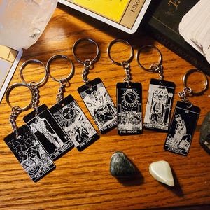 May include: A collection of black keychains featuring tarot card designs with silver-colored metal rings. The cards display intricate white illustrations and text, including "The Moon" and "The Sun". The keychains are arranged on a wooden surface.
