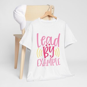 Peut inclure: T-shirt blanc avec l'inscription "Lead By Example" en lettres roses et jaunes. Le t-shirt est suspendu à un cintre en bois. Une casquette de baseball blanche repose sur le dossier d'une chaise en bois.