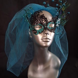 May include: A black and teal masquerade mask with a teal veil. The mask has a floral design with teal glitter accents. The mask is adorned with teal berries and peacock feathers. The veil is a sheer teal fabric.
