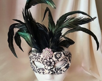 May include: A decorative masquerade mask with black feathers and a floral design. The mask features a cream-colored base with black floral patterns and a pink jeweled embellishment. The mask is topped with several black feathers.