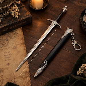 May include: A silver-toned metal sword with a black textured handle and a matching black scabbard featuring ornate silver filigree accents. The sword measures 22 cm in length and includes a silver keychain attachment on the scabbard.