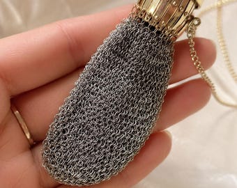 Könnte beinhalten: Ein kleiner, tropfenförmiger Beutel aus feinem, silberfarbenem Kettengeflecht. Die Oberseite des Beutels besteht aus einem zylindrischen, goldfarbenen Metallrahmen mit vertikalen Stäben und kleinen Nieten, befestigt an einer zarten goldfarbenen Kette.