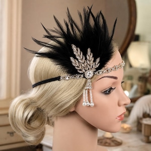 Pode incluir: Um acessório de cabeça com penas pretas e um design de folhas em metal dourado incrustado com strass transparentes. Inclui uma fina faixa elástica preta, um strass circular central e três contas brancas pendentes tipo pérola.