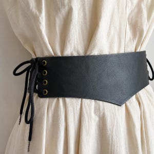 K&ouml;nnte beinhalten: Ein breiter, schwarzer G&uuml;rtel im Korsett-Stil mit strukturierter Oberfl&auml;che, messingfarbenen Metall&ouml;sen und schwarzer Schn&uuml;rung an den Seiten. Der G&uuml;rtel wird &uuml;ber einem hellen, gerafften Kleidungsst&uuml;ck getragen und betont die Taille.