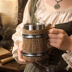 Pode incluir: Uma caneca cilíndrica com uma textura de grão de madeira castanha no exterior, acentuada por duas faixas horizontais em prata metálica. A caneca tem um interior liso em aço inoxidável e uma pega curva fixada na lateral do recipiente.