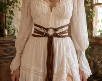 Könnte beinhalten: Ein cremefarbenes Kleid mit langen Ärmeln, Spitzenbesatz und gerafftem Oberteil, kombiniert mit einem braunen, mehrsträngigen Gürtel, der silberfarbene Mondsichel-Elemente und lange, herabhängende Fransen an der Taille aufweist.