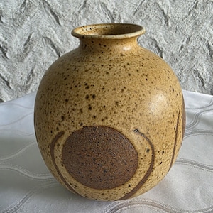 May include: A tan ceramic vase with a rounded body and a narrow neck. The vase has a speckled texture and a large, dark brown circular design on the front. The design is framed by curved lines. A decorative piece for the home.