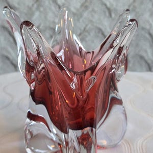 Puede incluir: Un cuenco decorativo de cristal transparente y rojo rubí. El cuenco tiene forma de estrella con bordes puntiagudos. El interior es de color rojo intenso, contrastando con el exterior transparente. El cuenco está sobre una superficie blanca.