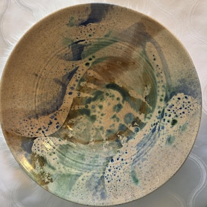 May include: A handmade ceramic plate with a speckled, textured surface. The plate features a blend of earthy tones, including beige, brown, and blue, with a splash design. The rim has a slightly raised edge.