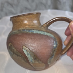 以下が含まれることがあります： 丸みを帯びたボディと湾曲したハンドルを備えた手作りのセラミックピッチャー。ピッチャーは、マルーン、ブルー、グリーンの縞模様のブラウン釉薬が特徴です。注ぎ口はわずかに傾斜しており、全体的なデザインは素朴な美学を示唆しています。