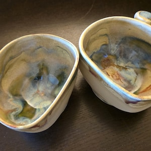 Op de afbeelding: Twee handgemaakte keramische kommen met een unieke, organische vorm. De binnenkant heeft een mix van groene, blauwe en bruine tinten, terwijl de buitenkant crèmewit is. Elke kom heeft een klein, afgerond handvat.