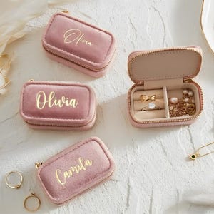 May include: Pink velvet jewellery travel cases, one open to display rings and necklaces. Two cases are closed with the name "Olivia" in gold script, and one with "Camila". Gold zips and jewellery are visible.