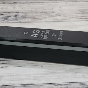 May include: A rectangular, black bar with a silver stripe and text that reads "AG Silver 107.868 50 Ounces". The top of the bar has the number "47" printed on it. The bar is sitting on a light wood surface.