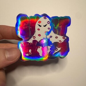 Puede incluir: Una pegatina holográfica que muestra un personaje fantasma blanco cubierto con patrones de labios rojos. El fantasma sostiene un teléfono de disco azul en su oreja. La palabra GHOST está impresa en letras grandes, oscuras y negritas.