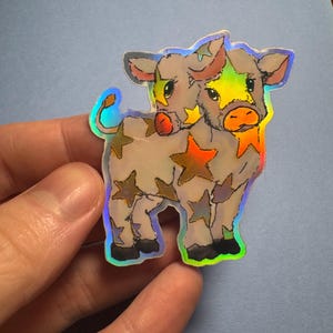 Pode incluir: Adesivo holográfico de uma vaca de duas cabeças com padrões de estrelas. A vaca tem um rosto colorido com as cores do arco-íris e é decorada com estrelas douradas, laranja e prateadas. O adesivo é segurado por uma mão sobre um fundo azul claro.