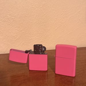 May include: Two pink metal lighters, one open and one closed. The lighters are on a wooden surface.