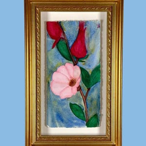 May include: A watercolour painting of a pink flower and two red buds on a stem with green leaves, set against a blue background. The artwork is displayed in a rectangular gold frame with a textured border and a white mat, creating a layered effect.