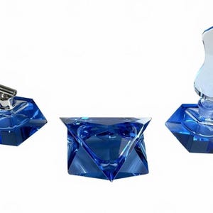May include: Three deep blue glass objects with faceted geometric bases. One piece features a silver-toned cylindrical stopper, one is a solid faceted block, and the third includes a tall, flame-shaped clear glass topper attached to the base.