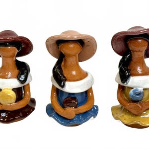 May include: Three ceramic figurines of women wearing hats and dresses. Each figure has a different coloured hat and dress, with one holding a yellow orb, one holding a brown square, and one holding a blue orb.