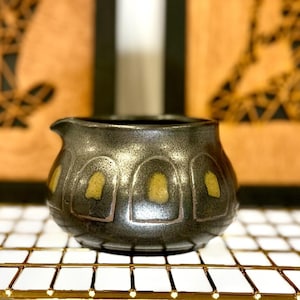 May include: A small, dark gray ceramic creamer with a spout and rounded body. The creamer features a series of arched, yellow-gold accents around its midsection. The background includes two framed artworks with abstract figures.
